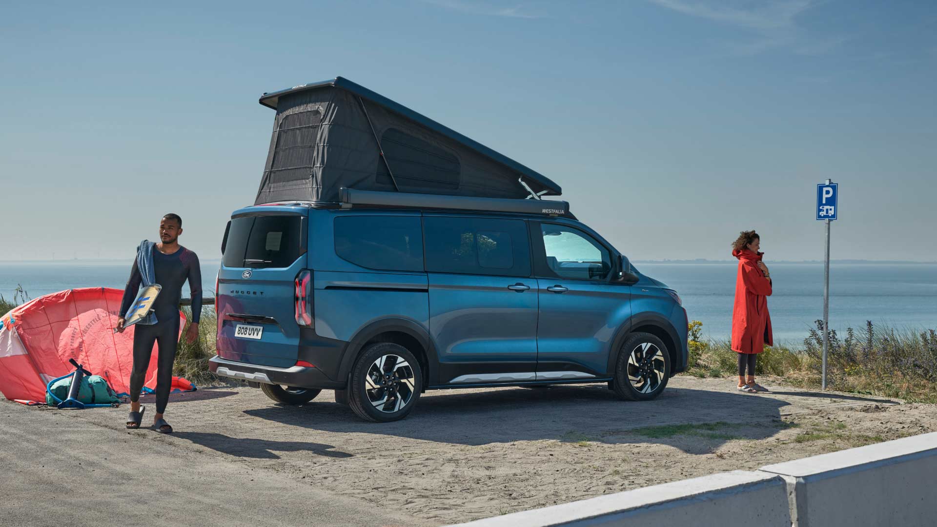 Un Ford Transit Custom bleu avec toit relevable est garé près de la mer. Deux personnes et une planche de surf sont à proximité.