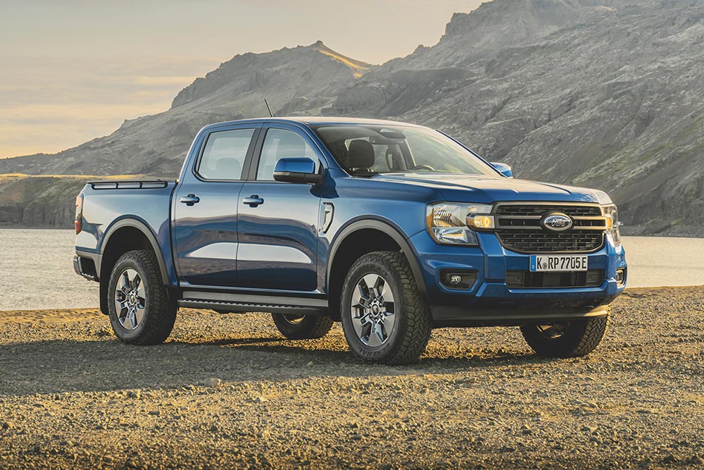 Un Ford Ranger bleu au bord d'un lac, avec en fond des montagnes escarpées et d'épais nuages.