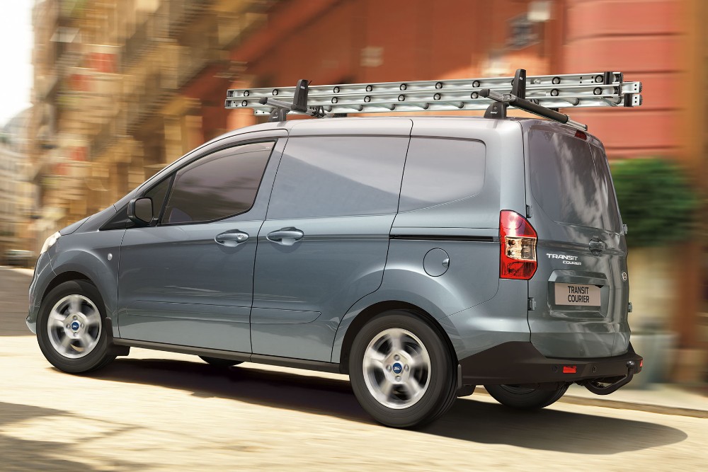 Ford Transit Courier, couleur bleu argenté, sur une route avec galerie de toit et échelle. Vue arrière aux trois quarts.