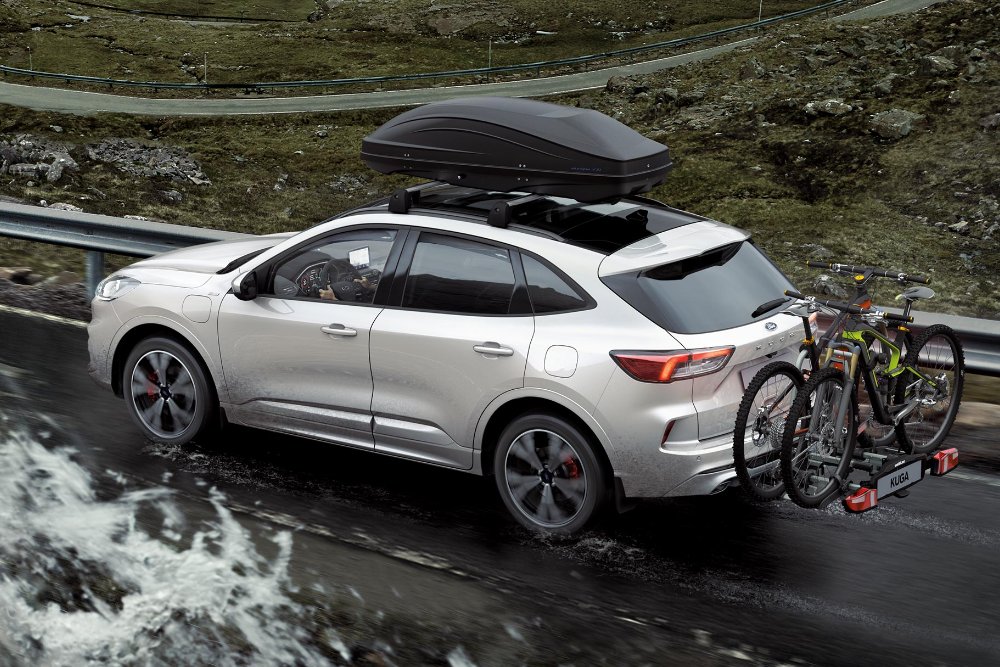 Ford Kuga, couleur argent, roulant sur une route de montagne avec un porte-vélos arrière et des vélos. Vue arrière aux trois quarts.