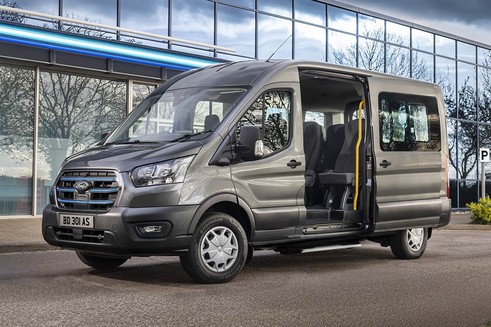 Vue latérale d'un Ford Pro Transit gris avec les portes ouvertes, garé devant un bâtiment moderne.
