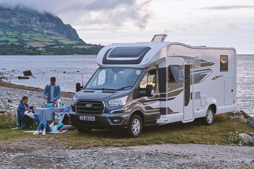 Un couple savourant des boissons dans des chaises de camping à côté d'un camping-car Ford Transit Custom gris avec un toit relevable.