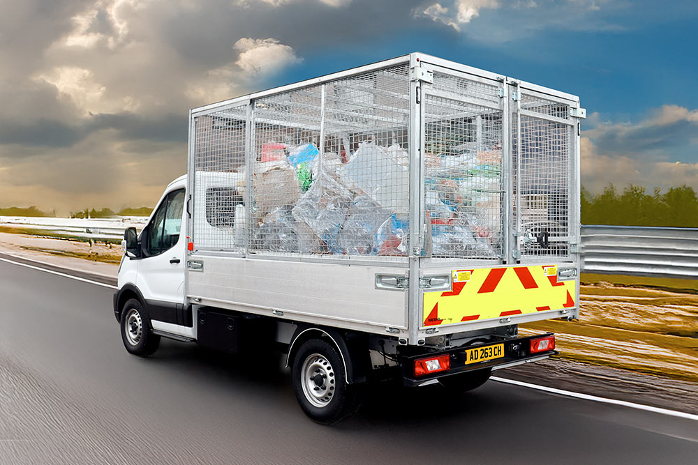 Vue arrière d'un camion benne à cage Ford Transit blanc, rempli de déchets, roulant sur une route.