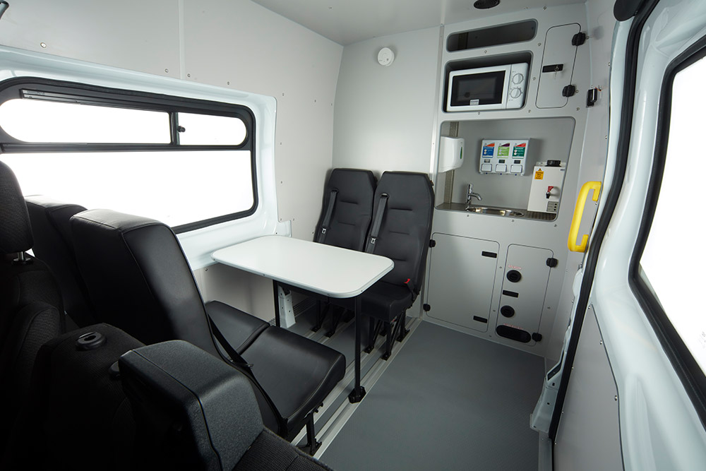 Intérieur d'une camionnette Ford, avec une table, des sièges noirs et une kitchenette compacte avec un micro-ondes.