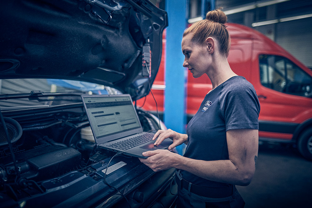 Un technicien Ford utilise un ordinateur portable pour diagnostiquer un véhicule, capot ouvert, dans un atelier.
