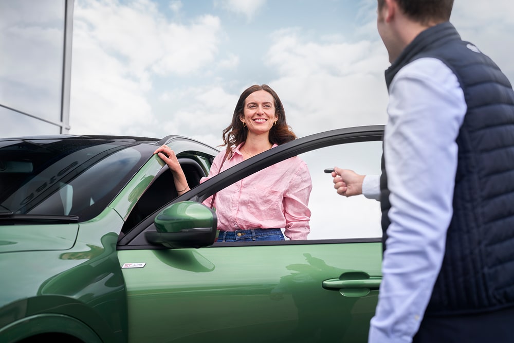 Une femme vêtue d’une chemise rose souriant à côté d’une voiture verte, avec un conseiller Ford qui lui remet ses clés de voiture.